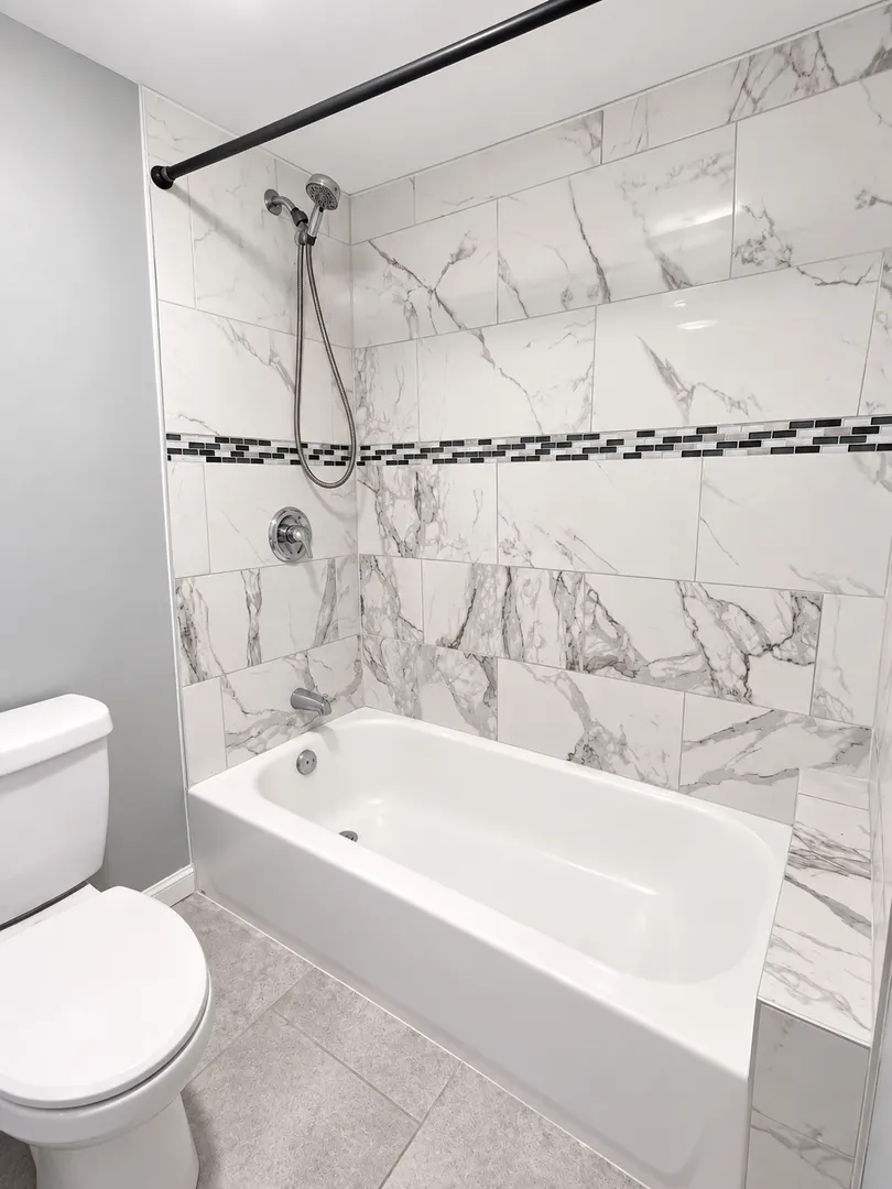 Bathtub with a white marble-look tile surround, a band of black and grey mosaic tile, a shower valve and a hand shower on a hose.