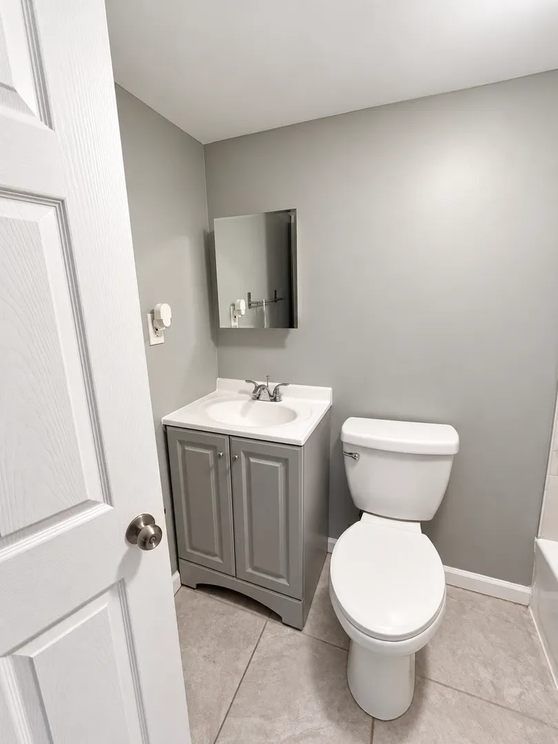 Bathroom with a grey vanity cabinet, white sink, mirrored medicine cabinet and a white toilet on a tile floor.