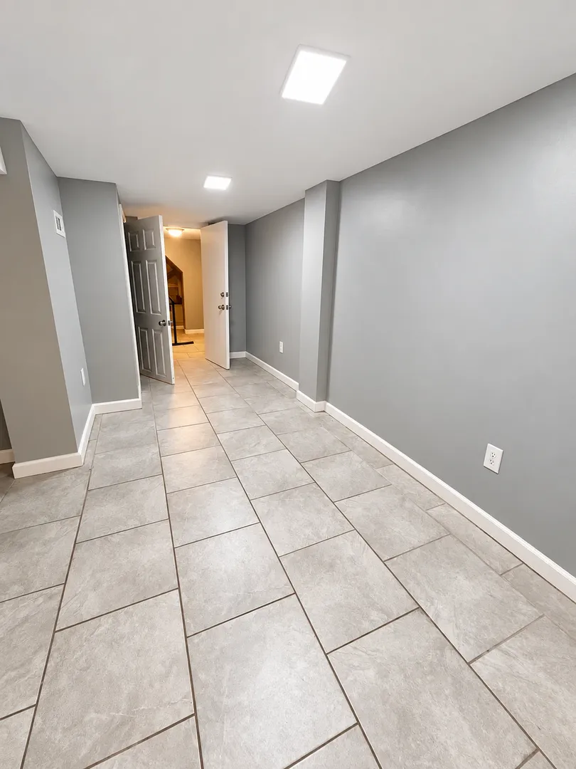 Long tiled corridor with grey walls and ceiling lights, leading to an open door and stairs.