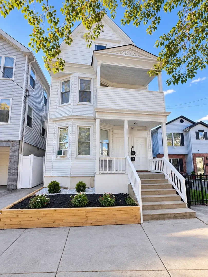Front of a white two-and-a-half storey multi-family house with a covered porch, upper balcony, front steps with white railings, and a raised timber planting bed along the sidewalk.