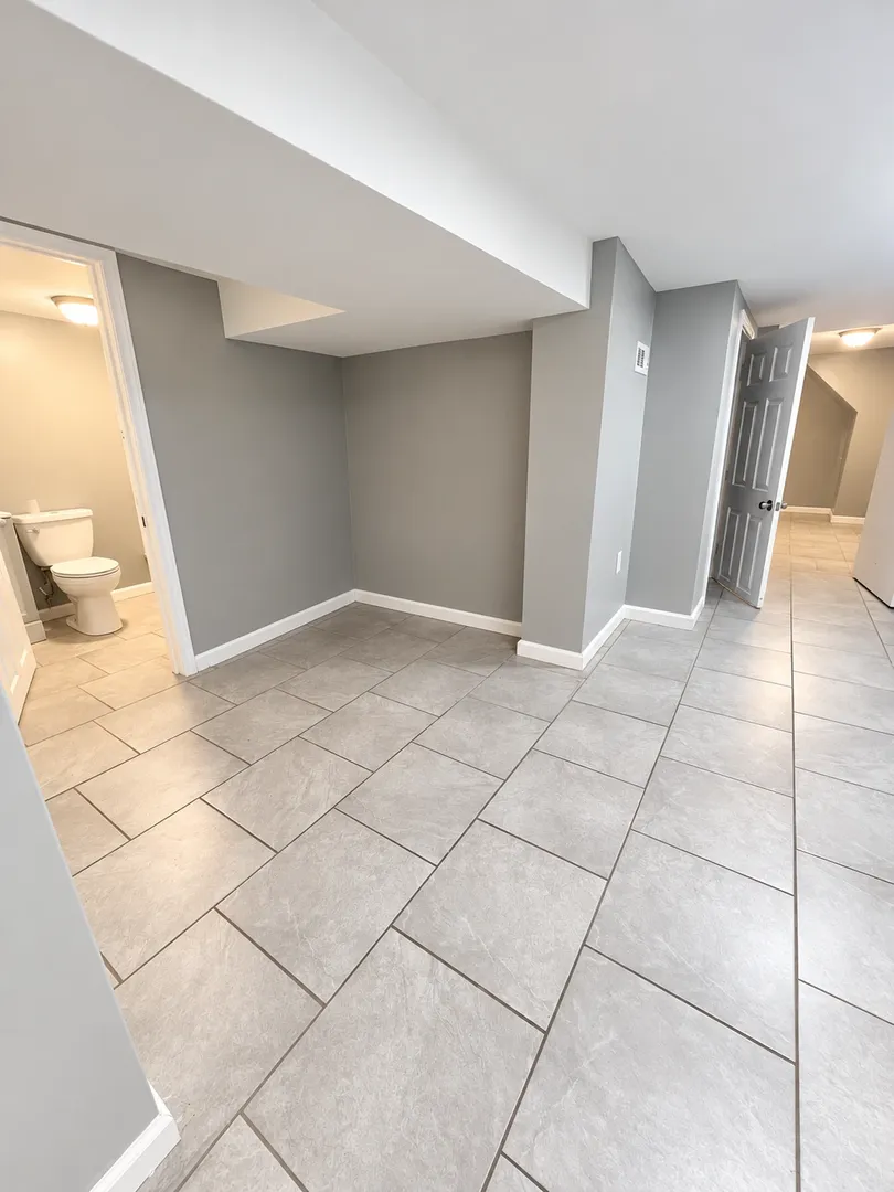 Hallway with grey walls and tile floor, a bathroom visible through an open doorway on the left and an open door ahead on the right.