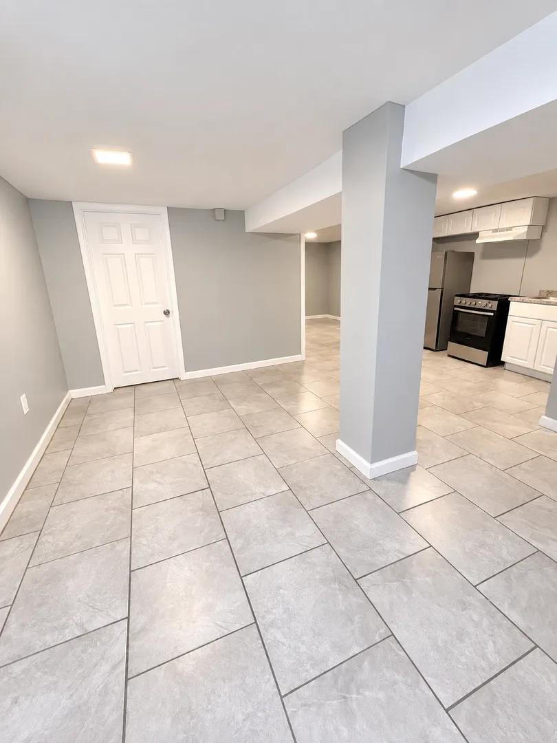 Open basement living area with grey walls, white trim, a boxed support column and large grey floor tiles, opening onto a kitchen with white cabinets.